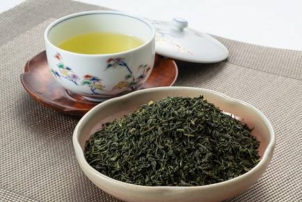 小皿に乾燥した深い緑色の茶葉が山盛りに盛られ、奥には木製コースターに載った色鮮やかな花柄の湯呑みに注がれた黄金色の緑茶が添えられた苣木茶を捉えたイメージ写真