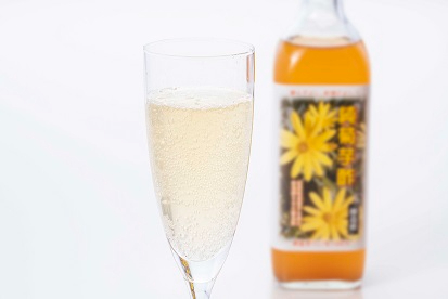 手前のフルートグラスに注がれた繊細な泡が立ち上る淡い黄金色の「菊芋酢」の炭酸割と奥には黄色い菊の花が描かれたラベルのボトルが配置されている爽やかな飲用スタイルを提案するイメージ写真