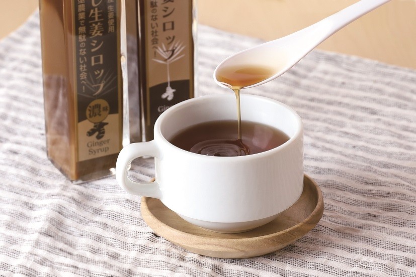 白いティーカップに注がれた茶褐色の飲み物に、白いスプーンからとろみのある琥珀色のシロップが静かに注がれている様子と、背景に「蒸し生姜シロップ」のボトルが2本並んでいる写真