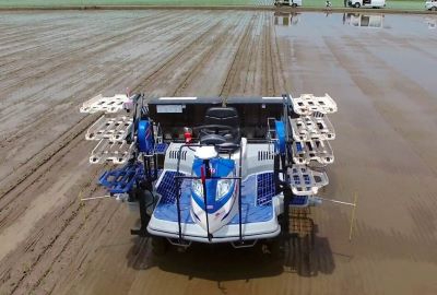 水田を走行する青と白の無人田植機を後方から俯瞰した視点で捉えており、機械の両脇には苗を載せるための予備苗台が左右に四段ずつ配置され、並行に整えられた泥の跡が奥へと続く晴天の下での作業風景の写真
