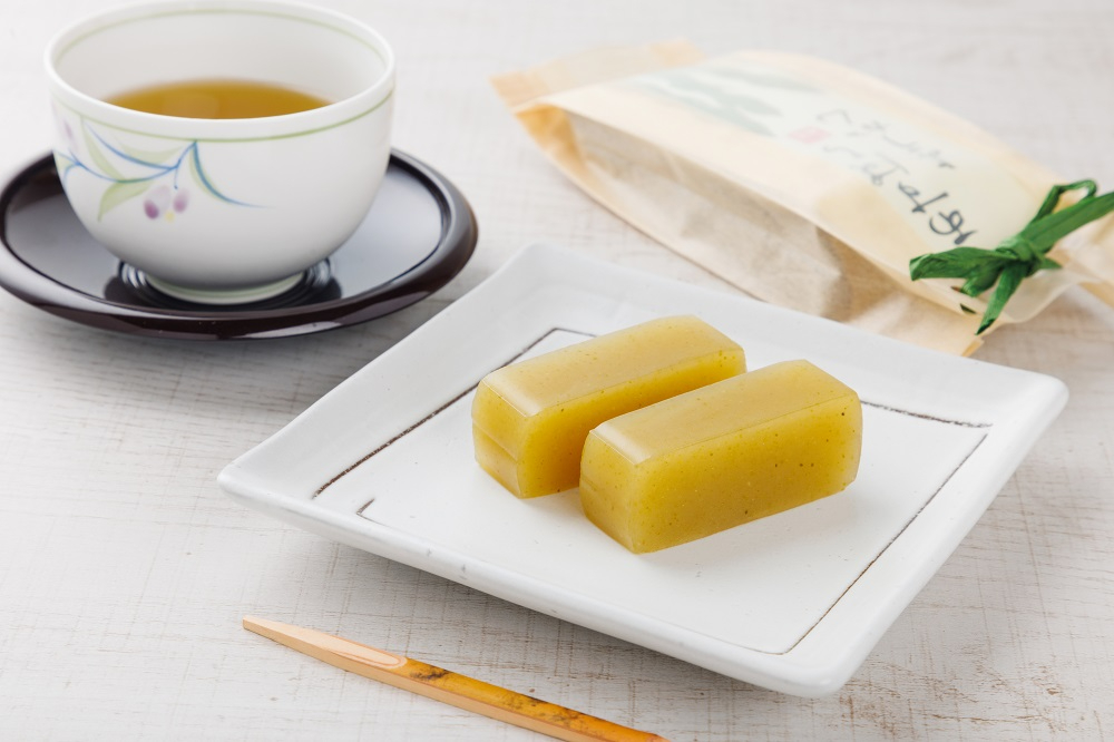 四角い白皿に一口大にカットされた淡い緑色の「アスパラ羊羹」が2個並び、奥にはお茶が入った白い湯呑みと個包装のパッケージが添えられたイメージ写真