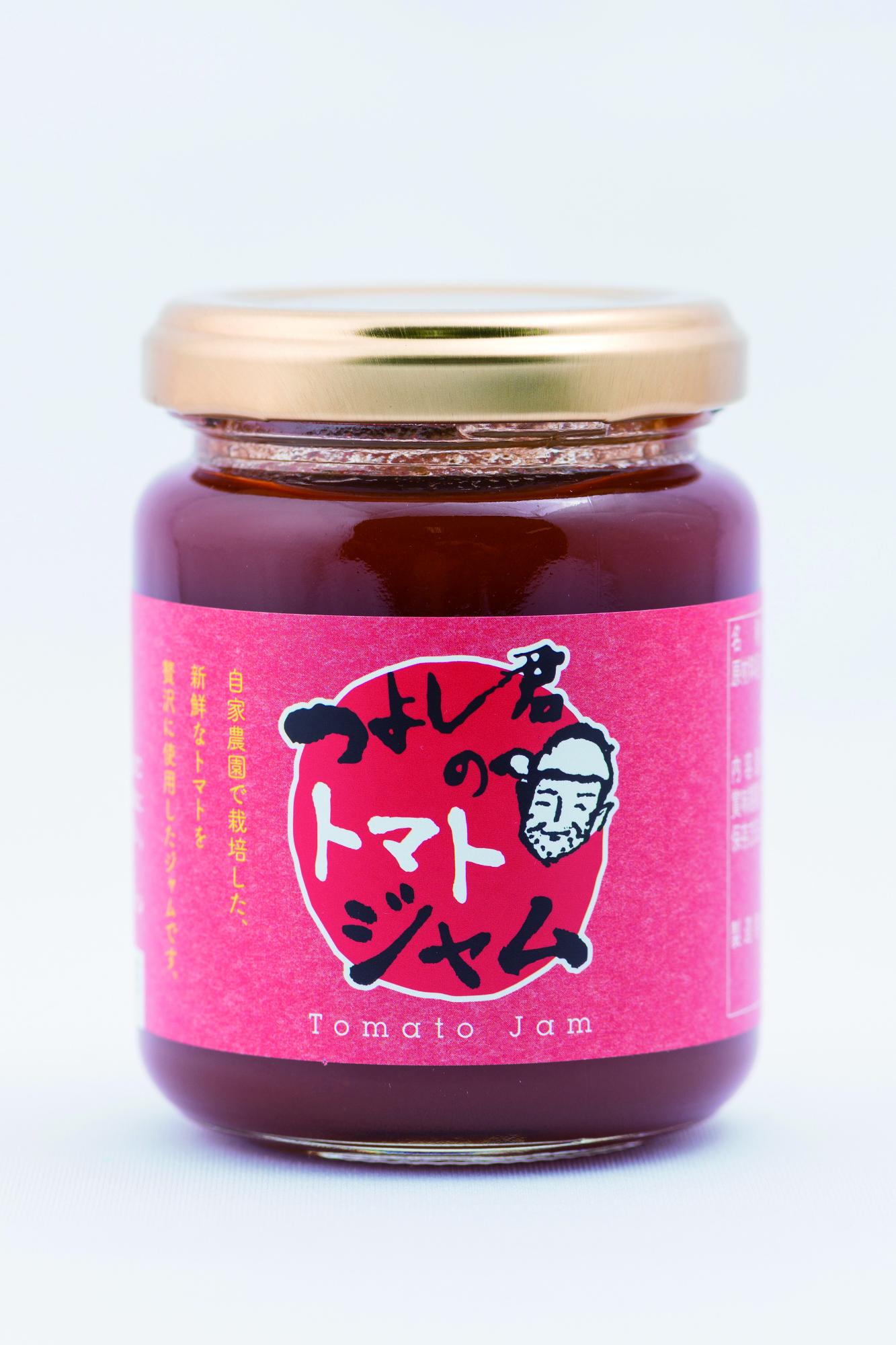 大きな赤い円の中に「つよし君のトマトジャム」という墨文字と男性の似顔絵がデザインされたラベルが貼られ、濃い赤色のジャムが詰まった金色の蓋付きガラス瓶の商品の写真