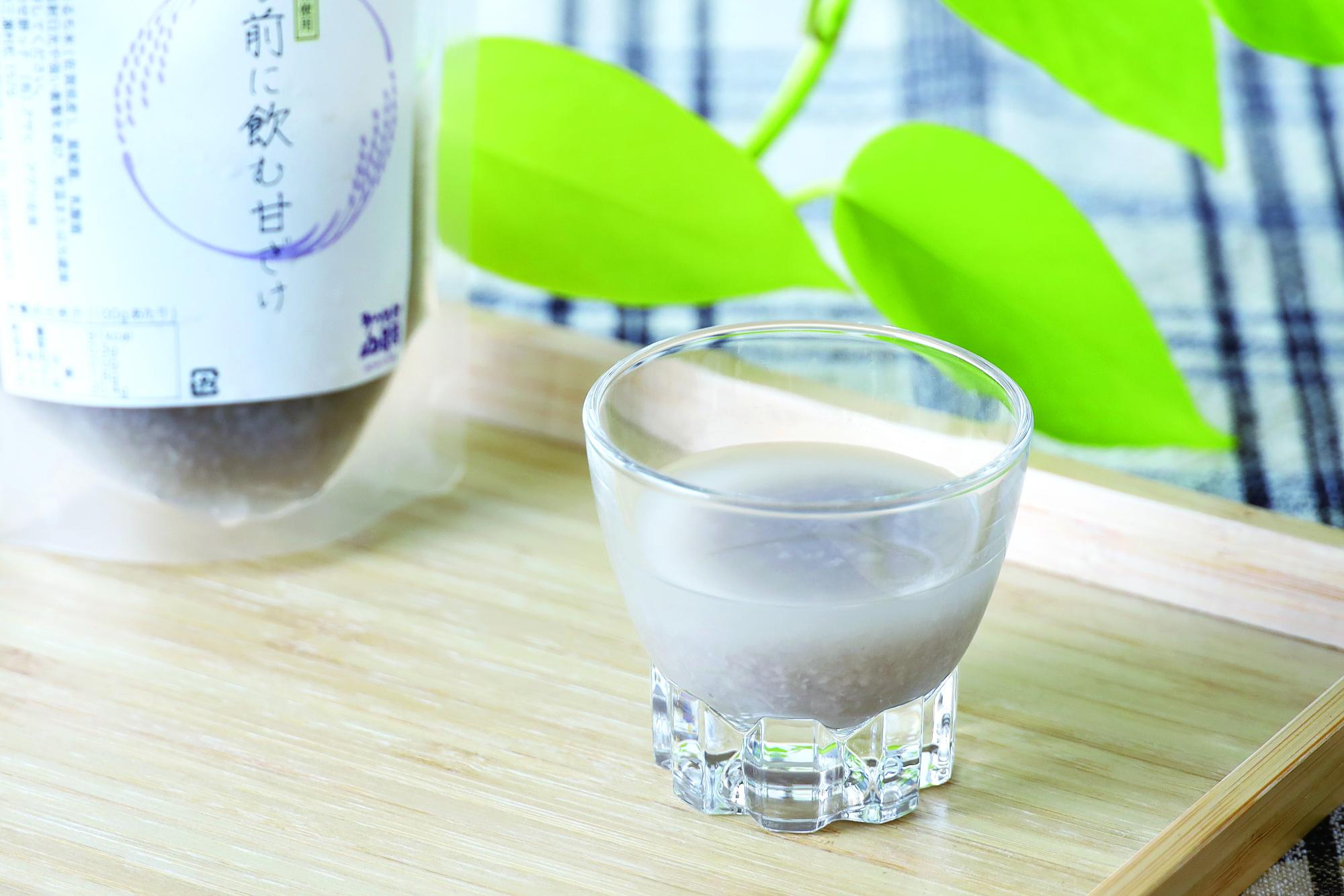 背景に鮮やかな緑色の葉が添えられ、木製トレイの上に商品パッケージと小さなグラスに注がれた白濁した「寝る前に飲む甘ざけ」が置かれている写真