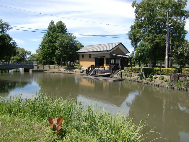 静かな池のほとりに建つ平屋の建物と、その周囲を囲む豊かな木々や草花、そして左側には小さな橋が架かっているボート乗り場の写真