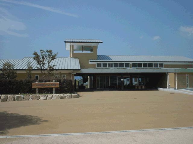 平屋建てのモダンな施設が広々とした舗装された地面の奥に佇み、建物の正面には木製の看板や植栽が整然と配置されている、清潔感のある四季のめぐみ館の外観写真