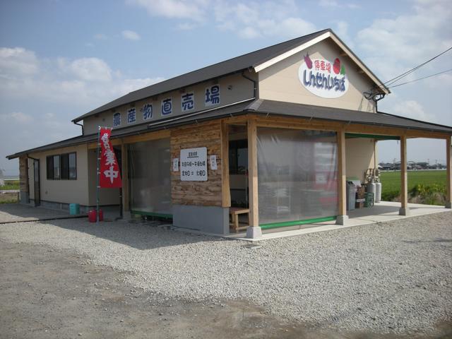 砂利が敷かれた広い敷地に建つ平屋の建物で、壁面に「農産物直売所」の文字や「しんせんいちば」というロゴ看板が掲げられ、入り口には赤い「営業中」ののぼり旗が立つ農産物販売所の外観を写した写真