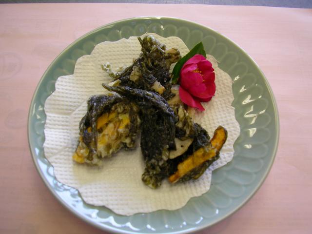 薄緑色の縁取りがある皿に敷かれた白いペーパーの上に、海苔と野菜が衣で揚げられたかき揚げが赤い花と盛り付けられた写真