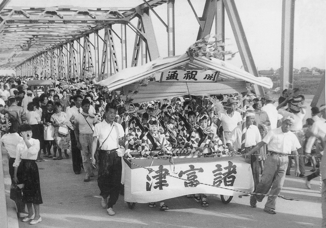 橋の上で多くの人々が山車を囲みながら賑やかな祭りの行列を進める活気ある様子を捉えたモノクロ写真