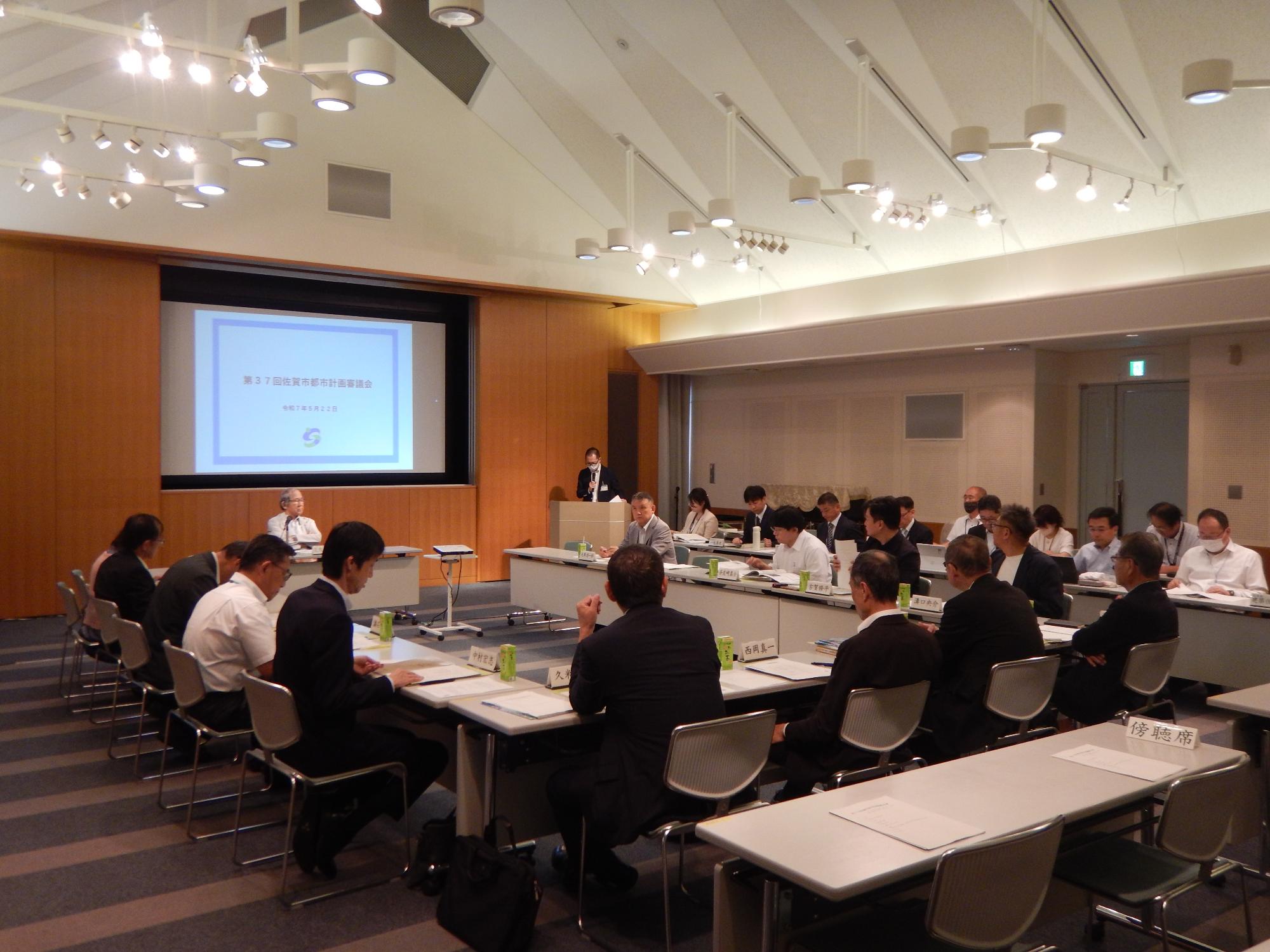 明るい会議室で、スーツ姿の多くの参加者がU字型に配置された長机に座り、前方に投影されたスクリーンを注視しながら資料を確認している会議の様子を捉えた写真