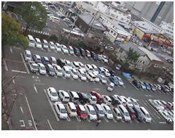 多数の白や黒、赤色の乗用車が整然と区画内に駐車され、建物や樹木に囲まれた屋外の広々とした平面駐車場の様子を上空から撮影した写真