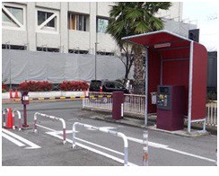 建物の前に設置された、赤い屋根付きの精算機と黄色と黒の縞模様のゲートバーが閉まっている駐車場の出入り口付近を、アスファルトの路面や背後の街路樹とともに正面から捉えた写真