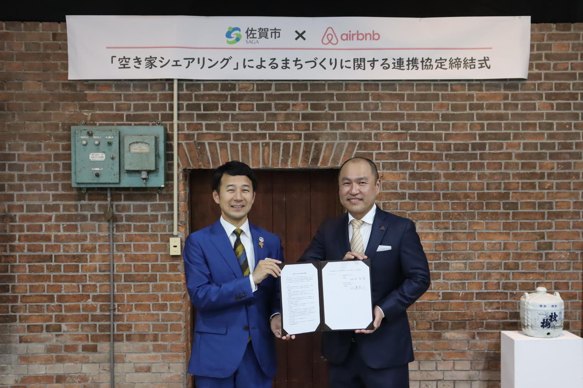 レンガ壁を背景に、上部に佐賀市とAirbnbのロゴと空き家シェアリングによるまちづくりに関する連携協定締結式の横断幕が掲げられ、スーツ姿の市長と田邉代表が協定書を持って記念撮影をしている公式調印式の写真