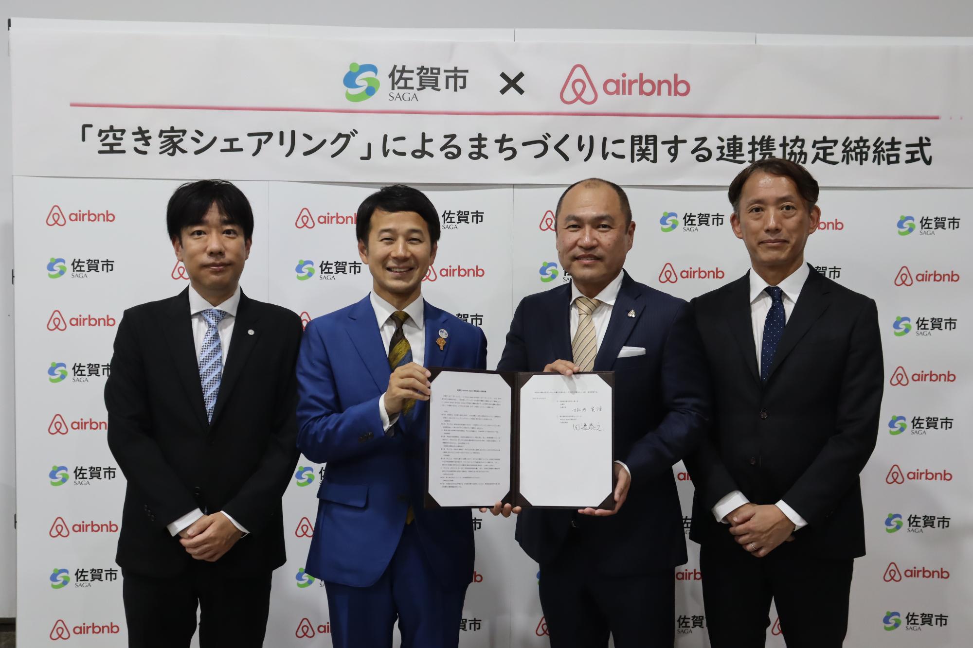 佐賀市とAirbnbの連携協定締結式で、スーツ姿の一丸都市戦略部長、坂井市長、田邉代表、谷口部長が署名文書を掲げ、両者のロゴ入り背景パネルの前に並んで記念撮影をしている写真