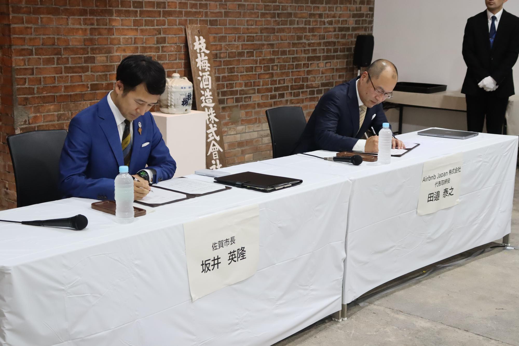 レンガ壁を背景に、白いテーブルクロスを掛けた長机に設けられた席で、佐賀市長坂井英隆とAirbnb Japan株式会社代表取締役田邉泰之が書類に署名する調印式の様子を撮影した写真