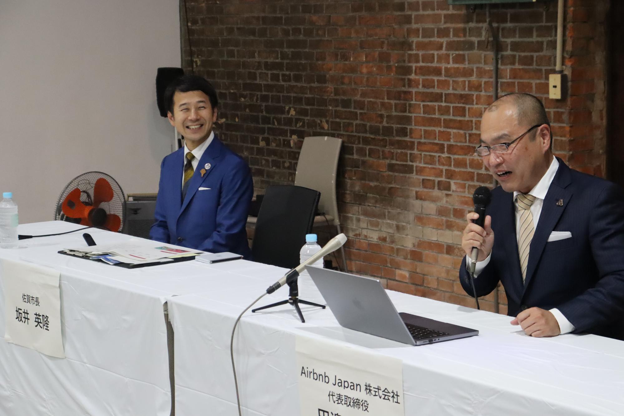 レンガ壁を背景に、白いテーブルクロスを掛けた長机に設けられた席で、佐賀市長坂井英隆とAirbnb Japan株式会社代表取締役田邉泰之の名札が並び、田邉代表がマイクを持って発言する様子を撮影した写真