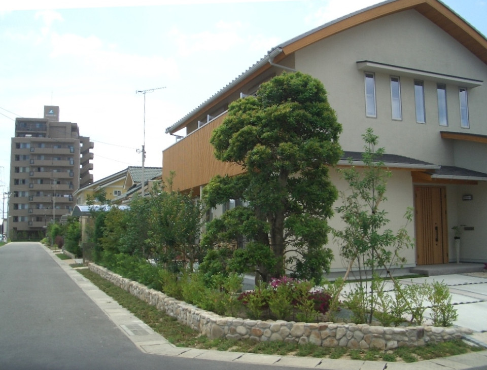 道路に沿って石造りの低い囲いがある花壇が設けられ、手入れの行き届いた植栽や中背の木々が新築の住宅を彩り、奥には並んで建つ家々や高いマンションが見える、美しく整えられた住宅街の街路樹と外構の風景を捉えた写真