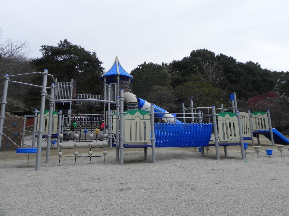 山や森を背景にした広大な砂地の公園に、とんがり屋根のタワーや青いトンネル滑り台、ネット登りや吊り橋などが複雑に組み合わさった、グレーと青を基調とする大規模でアスレチック要素の強い複合遊具ウインドライドを捉えた写真