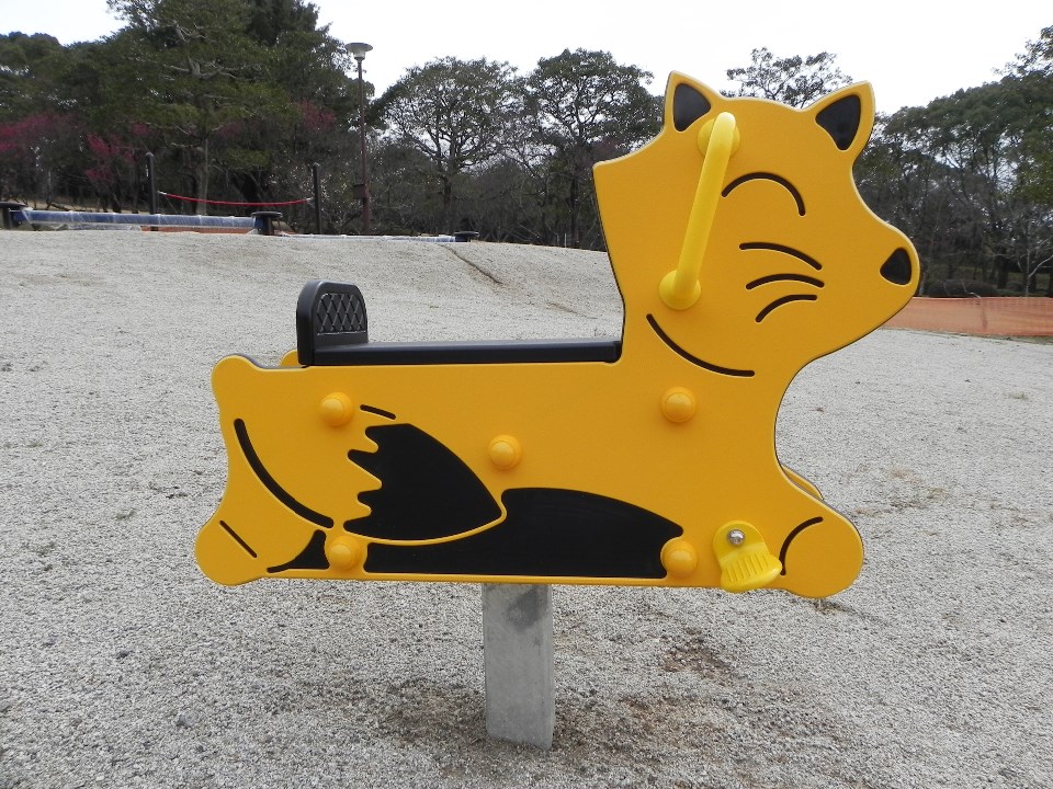 砂利が敷かれた公園の広場に設置された、ウィンクをする可愛らしいキツネの顔がデザインされた黄色いスプリング遊具で、黒い座席や手すりが付いた子供がまたがって遊ぶための乗り物遊具を横から捉えた写真