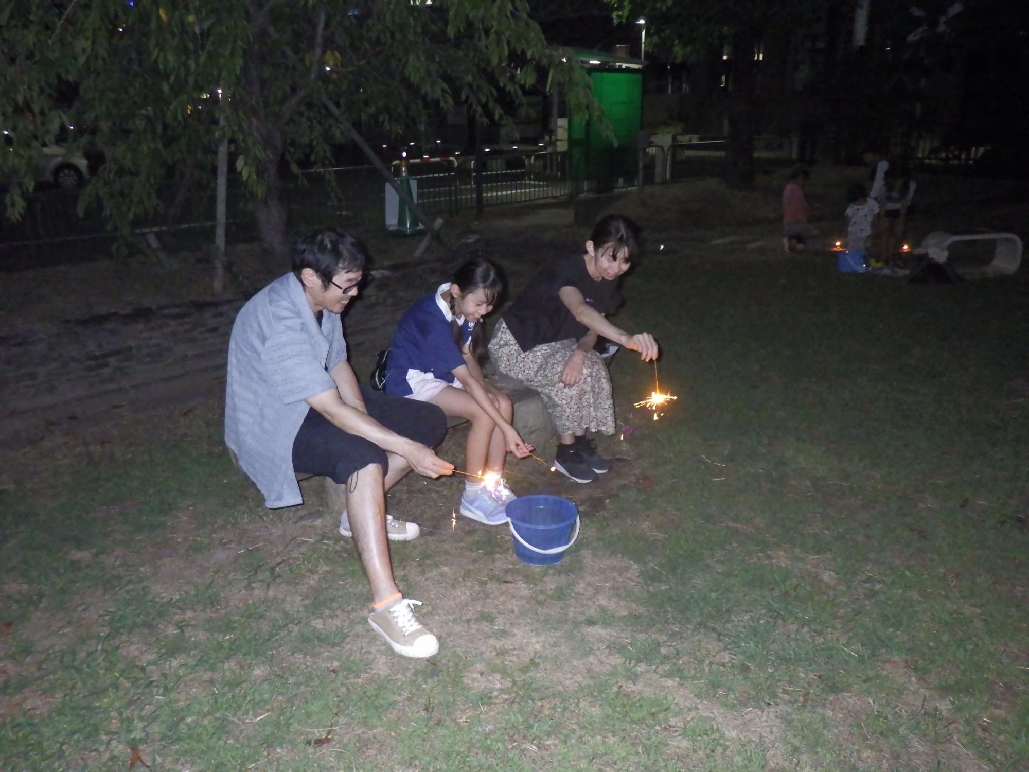 夜の公園の芝生に置かれたベンチに腰掛け親子連れと思われる3人が、足元のにバケツを置き、線香花火を楽しんでいる様子を写した写真