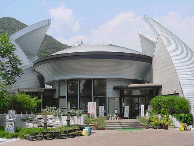 円盤のような形をした中央の屋根と左右に突き出した鋭い翼のような外壁が特徴的な近代的な建物の外観を、入り口前の階段や植栽を含めて正面から捉えた写真