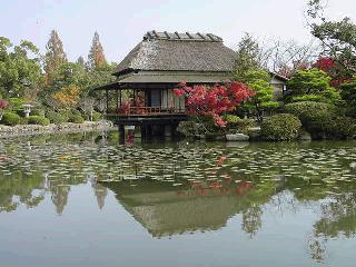 池のほとりに建つ茅葺き屋根の歴史的な茶室と、その姿を鏡のように映し出す水面、周囲を彩る鮮やかな紅葉や青々とした木々がある神野公園の写真