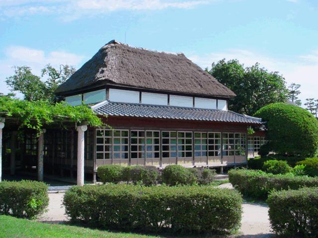 茅葺屋根の歴史的な建物と、その手前に広がる丁寧に手入れされた低い生垣や丸く刈り込まれた植栽、そして左側に続く藤棚のような棚構造がある神野のお茶屋の写真