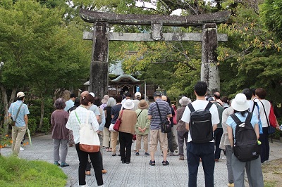 與止日女神社の鳥居の前で講師の説明を聞く参加者の様子。