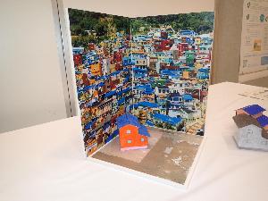ジオラマコーナーに設置するジオラマの見本の画像。色とりどりの家が並ぶまちなみの写真を背景素材として、オレンジ色の外壁・青色の屋根をした2階建ての家のジオラマが画像中心部に置かれている。