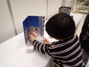 ジオラマで遊ぶ子どもの様子。白色や灰色のマンションが立ち並ぶ街の写真の前に、赤い屋根の家のジオラマを置こうとしている。
