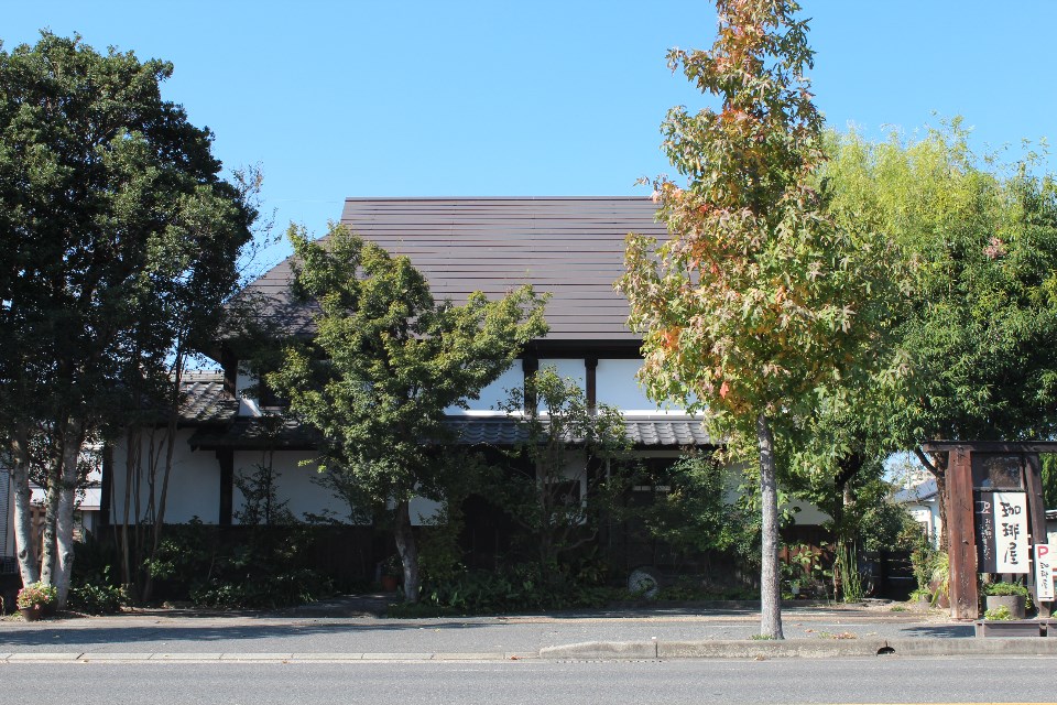 白壁と濃い茶色の木材を組み合わせた和風建物が道路沿いに建ち、前面には紅葉した木や緑の植栽が並び、右側には「珈琲屋」と書かれた木製看板が立つぎゃらりー喫茶 欒（まどい）の外観写真