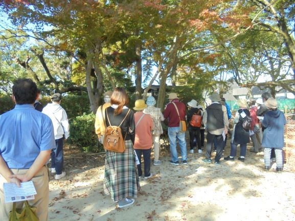 秋の木漏れ日が差し込む庭園で、紅葉し始めた木々の前に集まった参加者たちが、一列に並んでガイドの説明を熱心に聞きながら、歴史的な景観や自然の美しさを静かに堪能している様子の写真