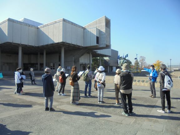 近代的なコンクリート建築が立ち並ぶ広々とした屋外広場において、鮮やかな青いジャンパーを着用したガイドが前方に立ち、資料を手にしながら建物を指し示す様子を熱心に見守る、等間隔に配置された参加者たちの姿を捉えた写真