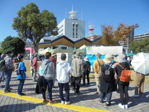 青空が広がる賑やかな屋外広場で、白いテントや特徴的な屋根の建物が並ぶイベント会場のような広場を訪れた十数人の参加者たちが、一列に並んで前方のガイドの話を熱心に聞いている様子を捉えた写真