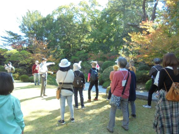 秋の陽光が降り注ぐ鮮やかな紅葉に包まれた庭園で、ガイドが腕を伸ばして前方を指し示しながら解説を行い、それを十数人の参加者が静かに聞き入っている様子の写真