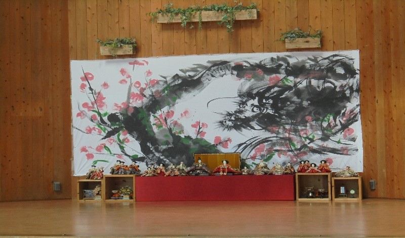 舞台上に桜を描いた大きな墨絵が掲げられ、その前に雛人形が整然と並ぶ展示の写真