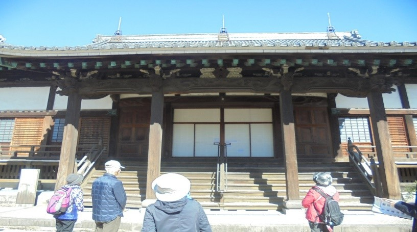 木造の本堂前で数人の参拝客が屋根の装飾や柱を眺めながら静かに佇む様子を捉えた写真