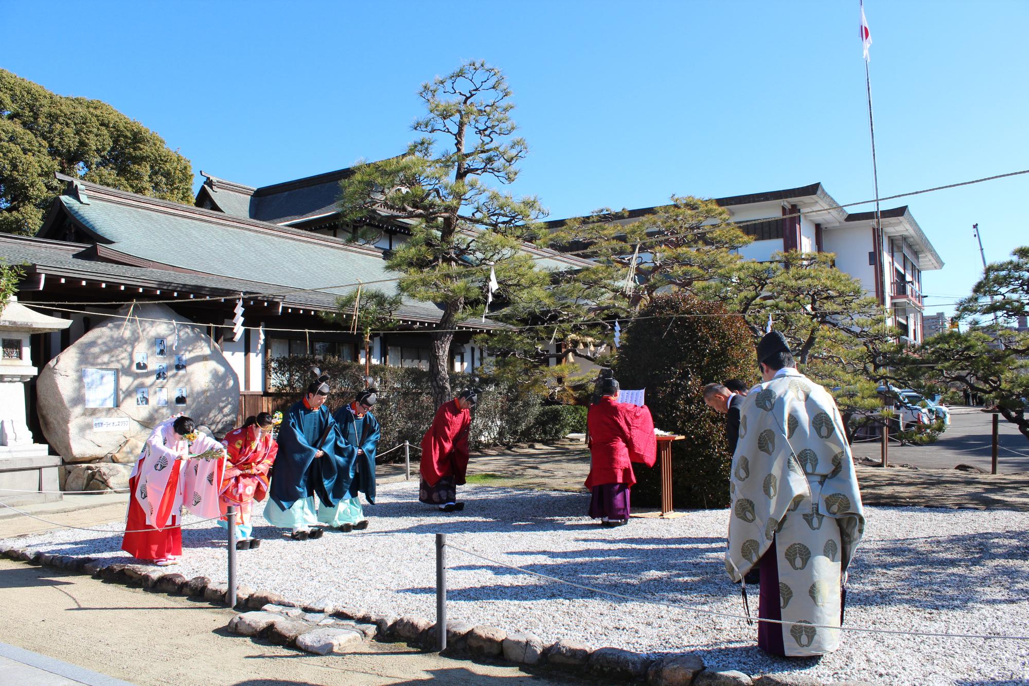 和装の人々が神社境内の白砂の庭で儀式のように整列し、社殿や松が背景に広がる写真