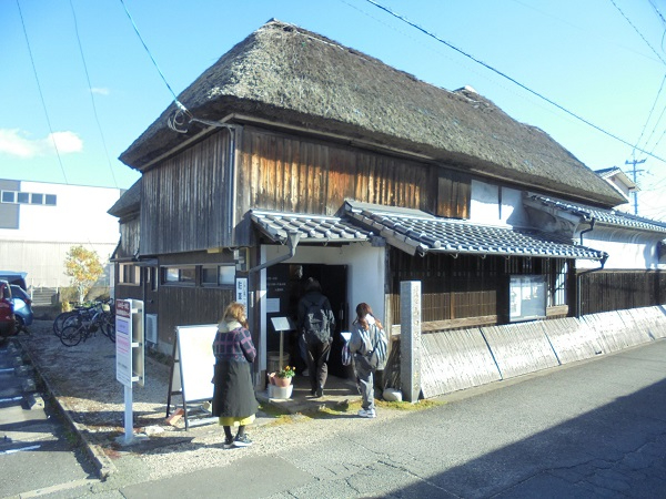 抜けるような青空の下、重厚な茅葺き屋根が特徴的な山口亮一旧宅の入り口に、リュックを背負った数名の見学者たちが建物の歴史を感じながら静かに入場しようとしている、冬の日の穏やかな街歩きイベントのワンシーンを捉えた写真