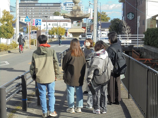 冬の柔らかな日差しが降り注ぐ中、歴史を感じさせる大きな石灯籠を囲むように集まった若者たちが、街の史跡や文化について熱心に意見を交わしながらフィールドワークを楽しんでいる写真