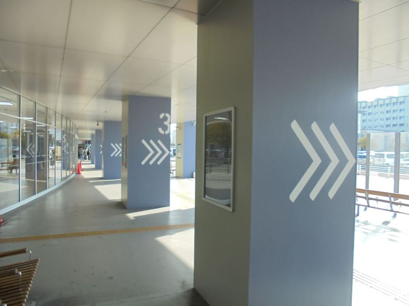 佐賀駅バスセンターのガラス張りの通路に並ぶ柱へ白い矢印標示が描かれ、光が差し込む駅前の乗り場通路を示す写真