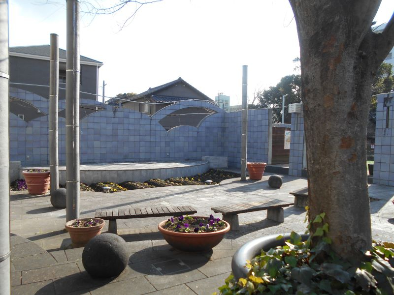 花壇やベンチが配置された小広場で青い装飾壁と木々が柔らかな日差しに照らされる唐人町まちかど広場の写真