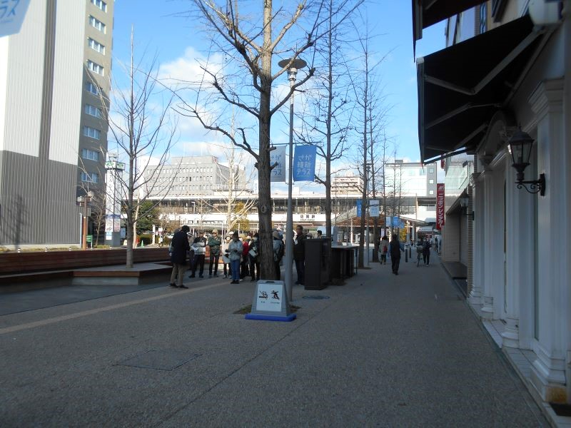 駅前の歩道沿いに並ぶ若木と商業施設を背景に、数人の来訪者が広場付近で集まり行き交う様子を収めた写真
