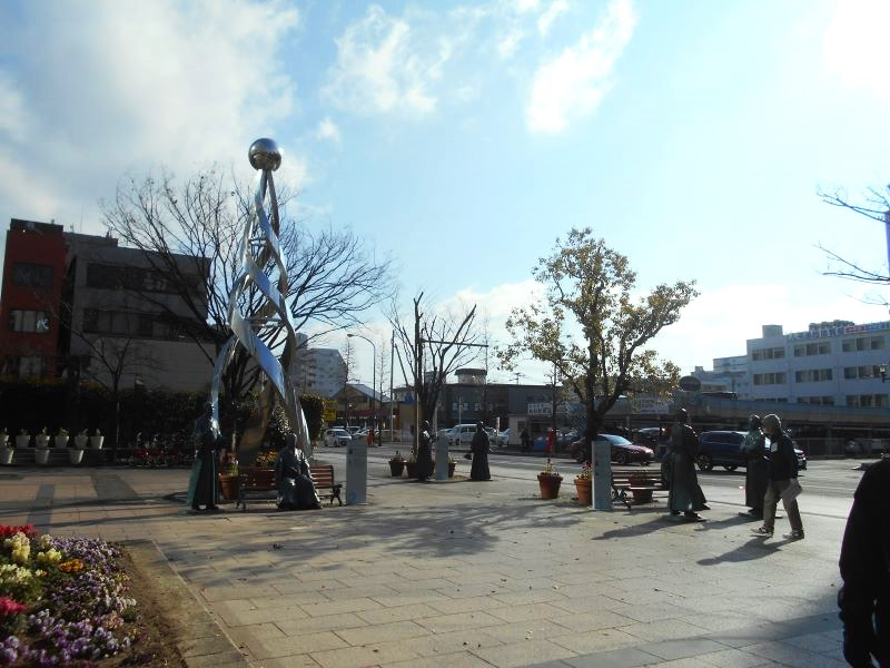 駅前まちかど広場で銀色の抽象的モニュメントと花壇が並ぶ中、通行人が冬の陽射しを受けながら行き交う様子を捉えた写真