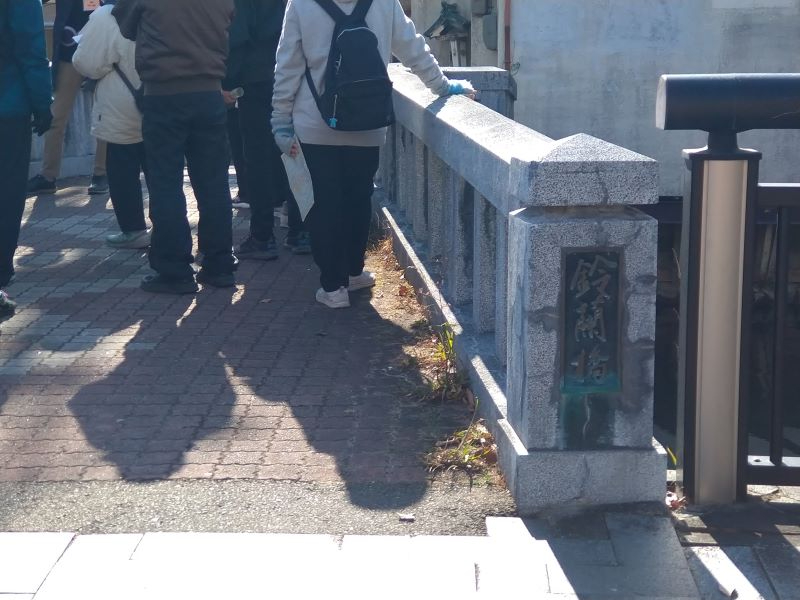 石造りの欄干に「鈴蘭橋」の銘板が付いた橋上で参加者が集まり説明を聞く様子を捉えた写真