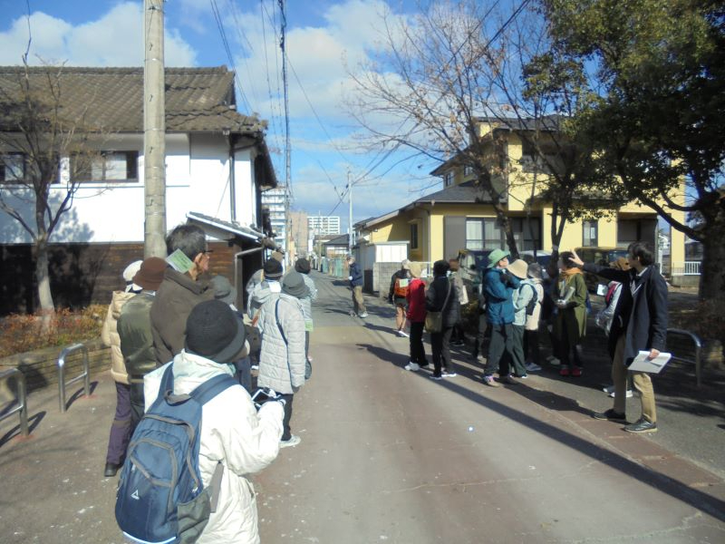 冬晴れの細い生活道路で参加者たちが左右に分かれて立ち止まり、周囲の住宅街を眺めながら説明を聞く様子を捉えた写真