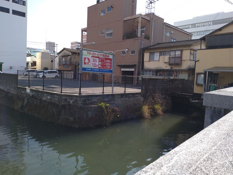 河沿いに石積みの護岸と古い建物が並び、駐車場の看板が立つ一角を穏やかな水面越しに捉えた写真
