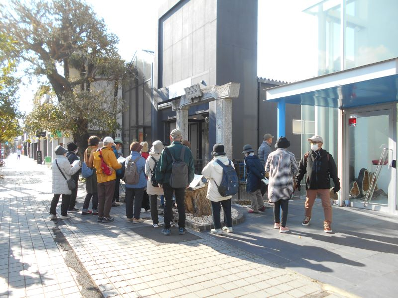 唐人町商店街の街路沿いの石製門柱前に集まった一団が説明を聞きながら建物周辺を見学している様子を捉えた写真