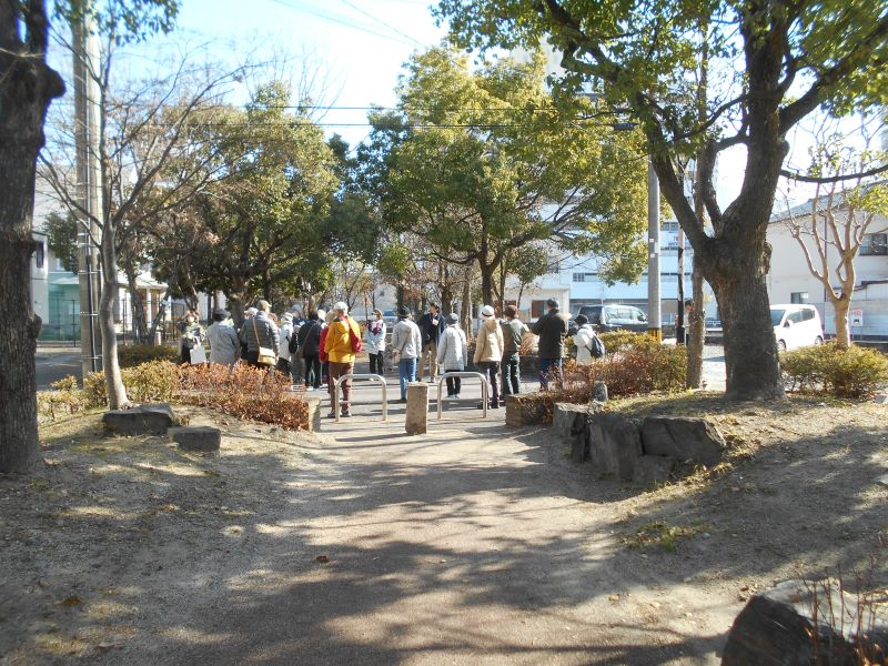 木々が並ぶ細い園路の先で大勢の参加者が集まり、日差しの差す公園内を散策しながら説明を聞く様子を収めた写真