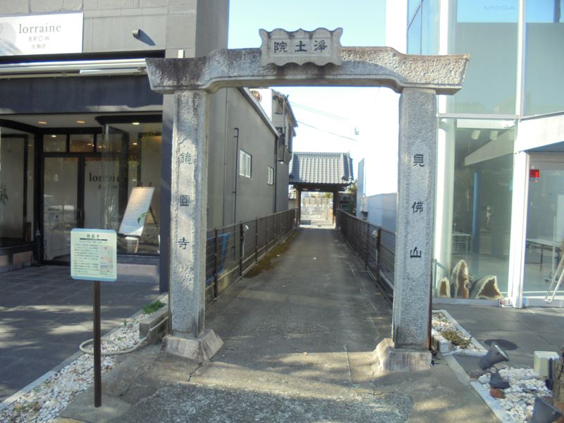 石造りの門柱が並ぶ細い参道の奥に見佛山鏡圓寺の屋根が見え、周囲の現代的建物と対比して静かな佇まいを示す写真