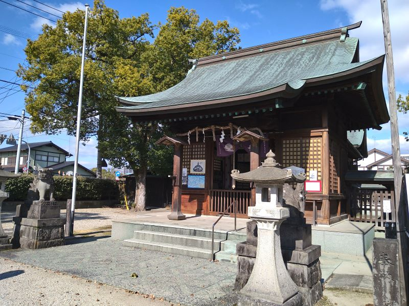 木製の拝殿が緑青色の屋根と石灯籠に囲まれて立ち、境内の木々と澄んだ青空が神社の静かな佇まいを際立たせる写真